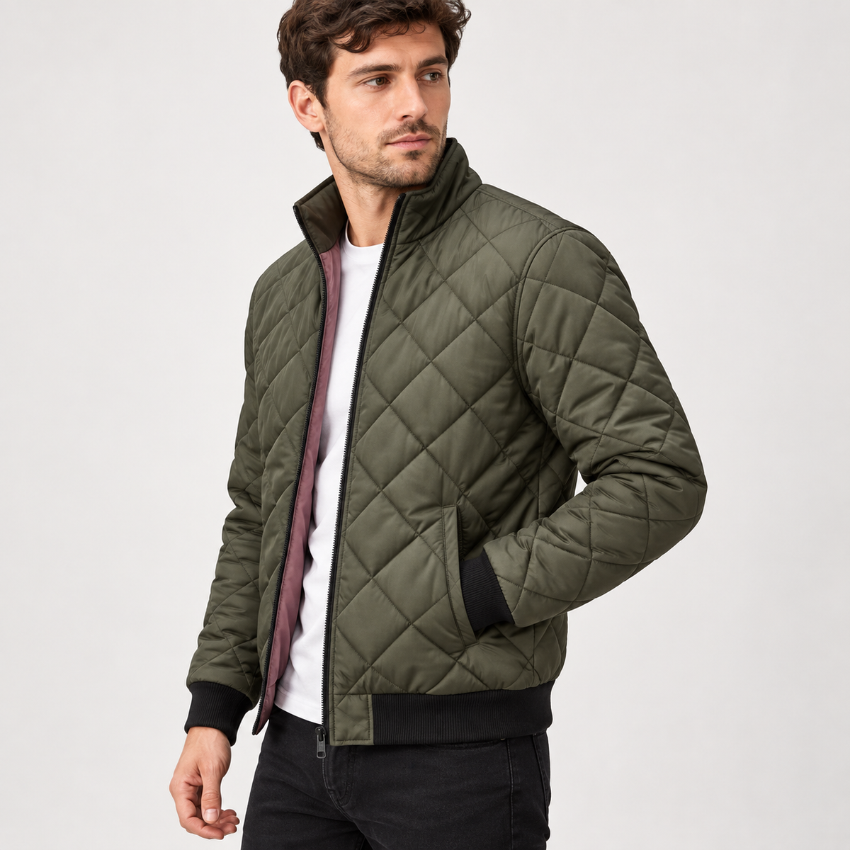 A man wears an olive green diamond-quilted bomber jacket with a black ribbed collar, cuffs and hem, open over a white t-shirt.
