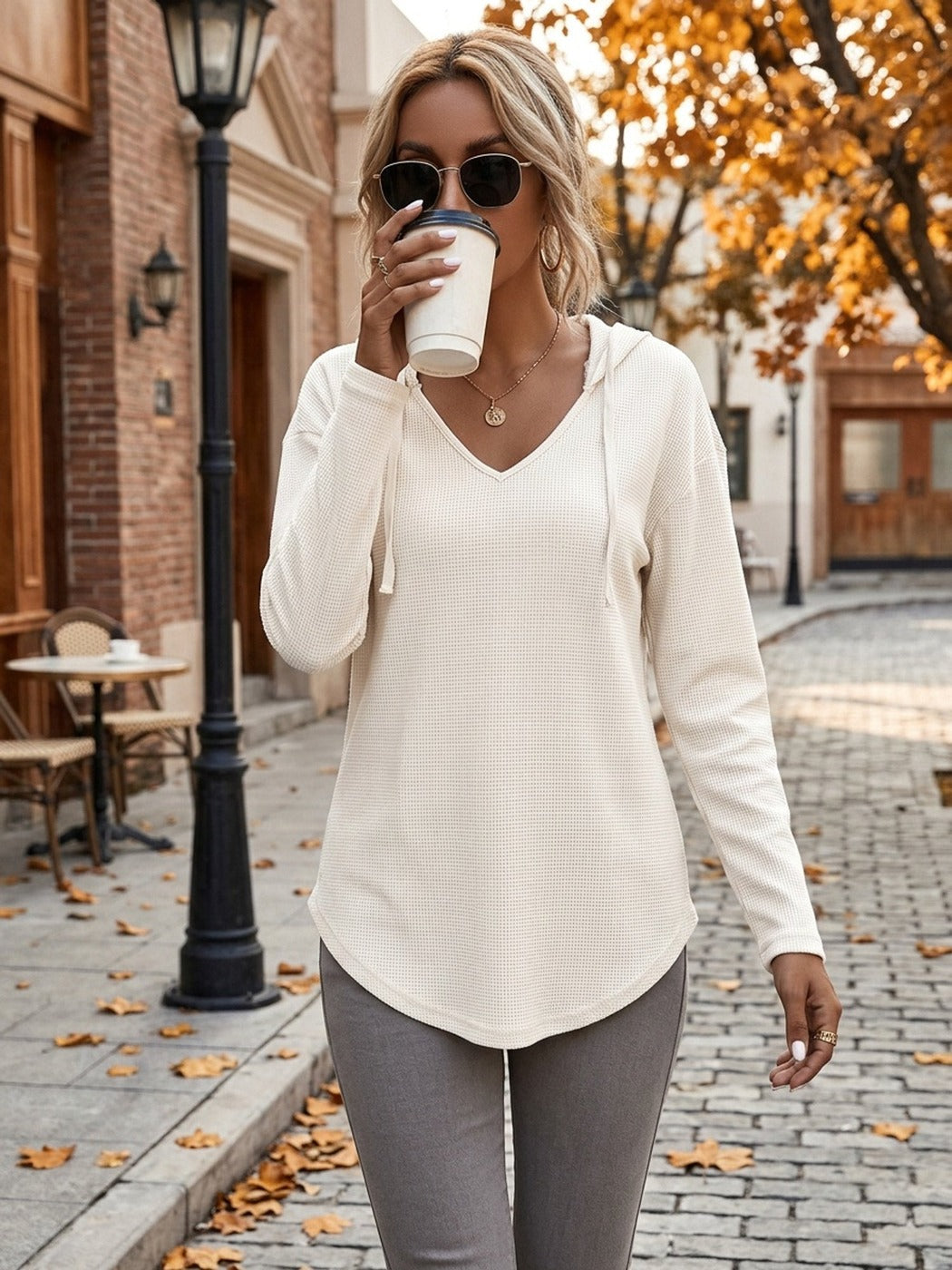 Vrouw in een gebroken witte hoodie met een grijze legging, koffie drinkend in een herfstige straat | Boutique Moda