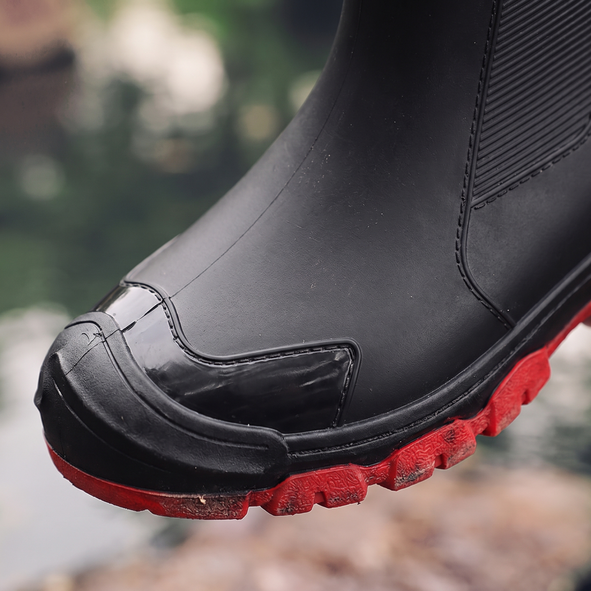 A pair of black ankle rain boots with red soles, one worn on a foot and one held by a hand, near water, close-up view