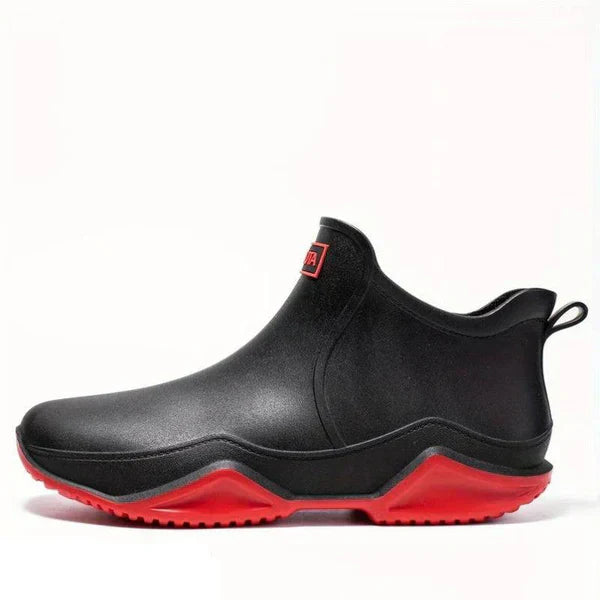 A black ankle-height rubber rain boot with a chunky red sole and a rear pull loop, shown in side profile on a white background.