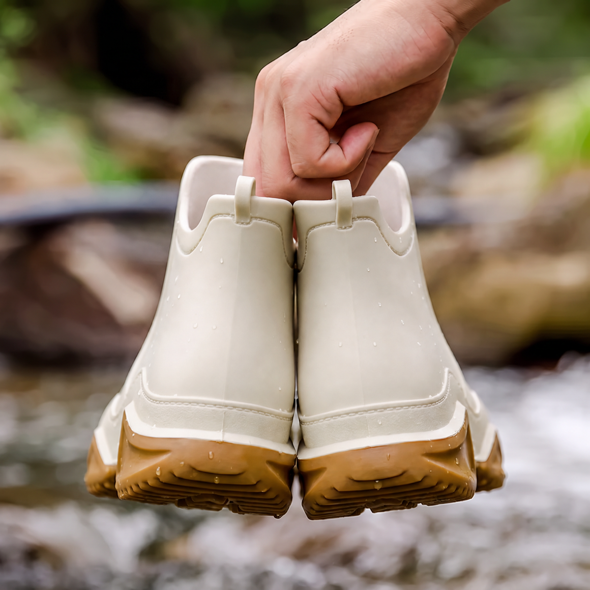 A hand holds a pair of cream ankle rain boots with tan soles above a rocky stream outdoors, back view