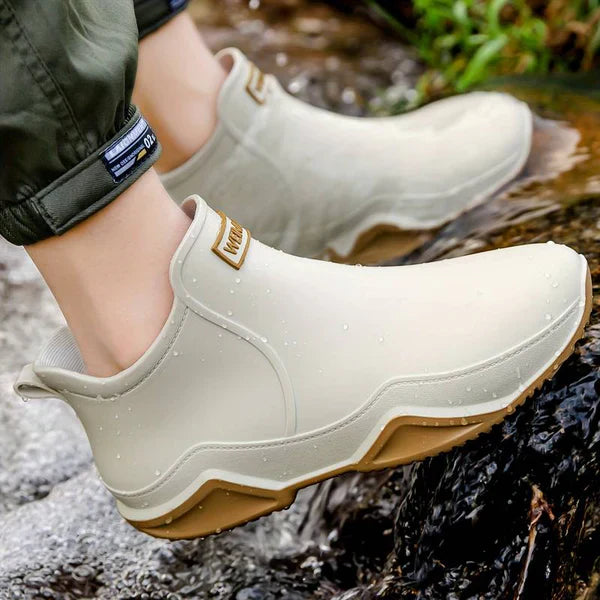 Cropped lifestyle shot of legs in green cargo trousers wearing cream ankle rain boots with tan soles, standing on wet rocks by water outdoors.