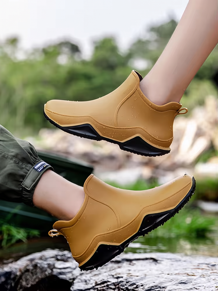 Lifestyle photo of a pair of mustard yellow ankle rain boots with black soles, worn on bare lower legs and lifted mid air outdoors beside water and rocks, with no face in frame.