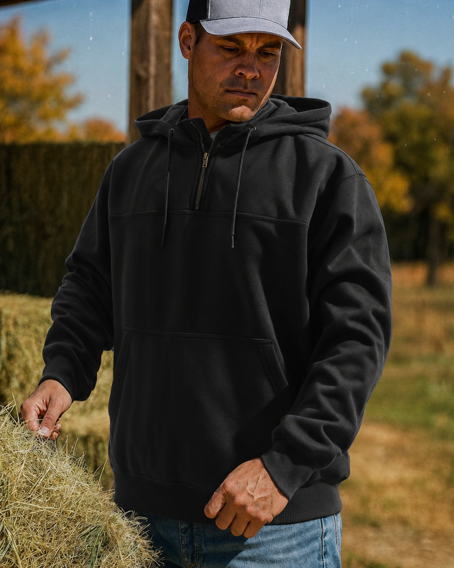 A man wearing a black quarter-zip hooded sweatshirt with a kangaroo pocket stands outdoors near hay bales.