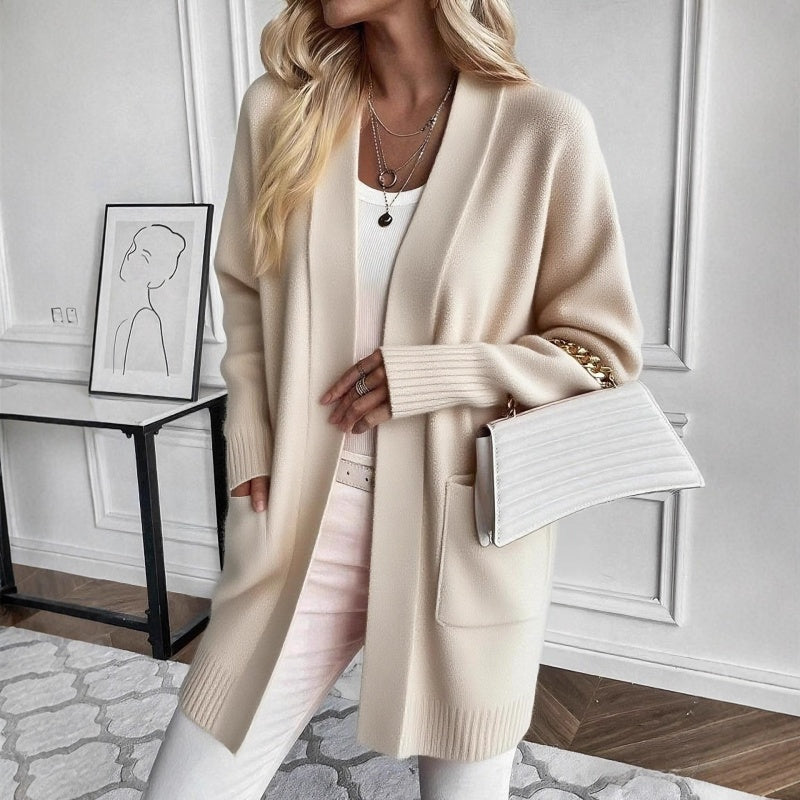 A woman wears a long beige open-front cardigan with ribbed cuffs and a front pocket, carrying a white handbag.