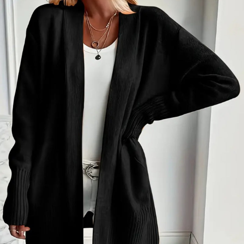 A woman wears an open black ribbed-trim long cardigan over a white top, with layered necklaces visible.