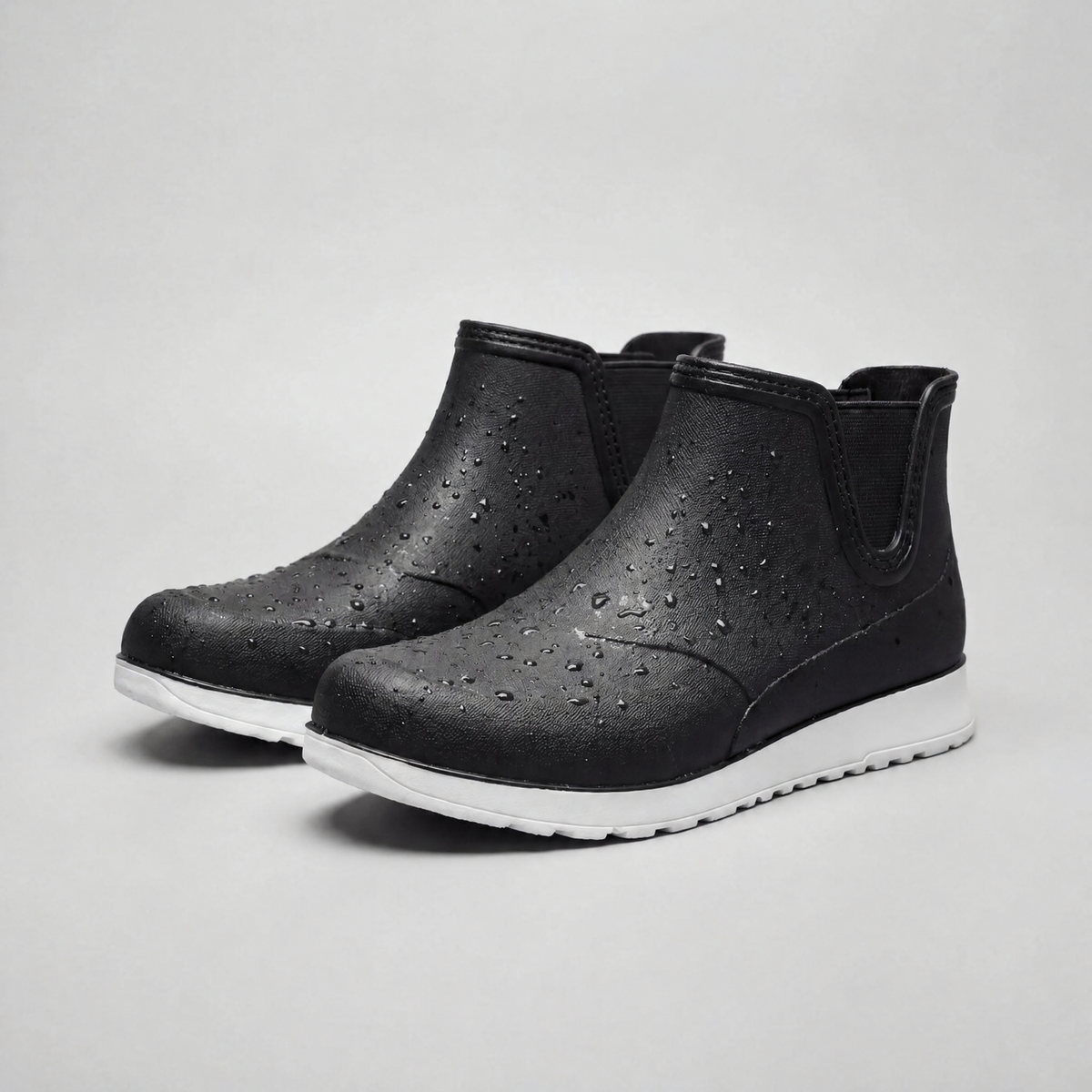 A pair of black rubber ankle boots with water droplets on the surface, white chunky soles, and elastic side panels, shown against a plain grey background.