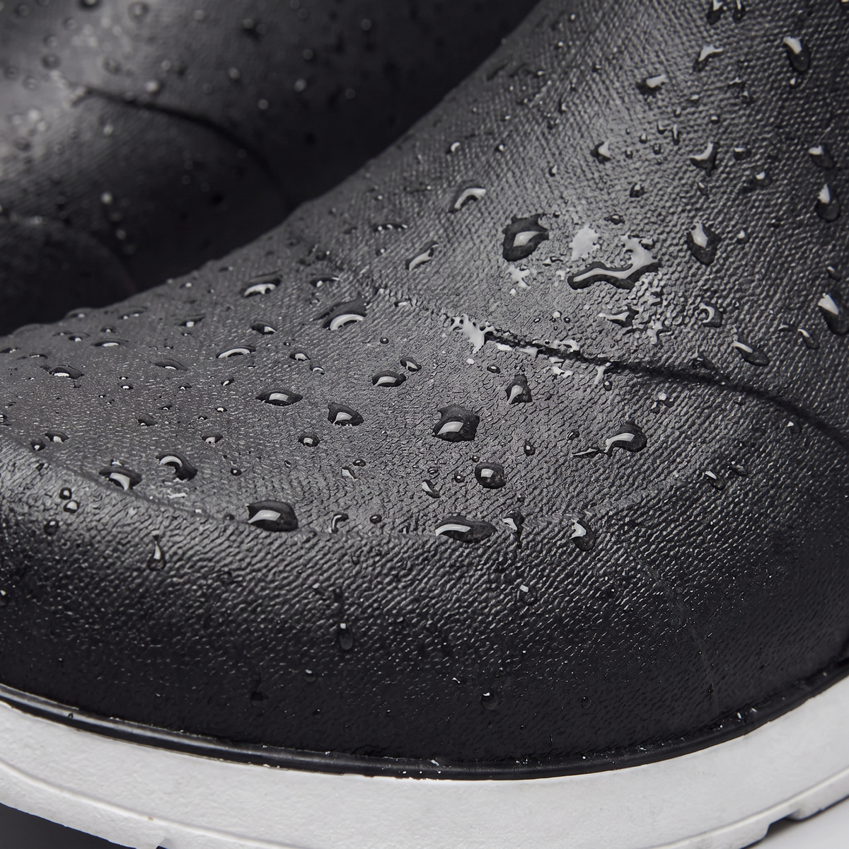 A pair of black rubber ankle boots with water droplets on the surface, white chunky soles, and elastic side panels, shown against a plain grey background, close-up view