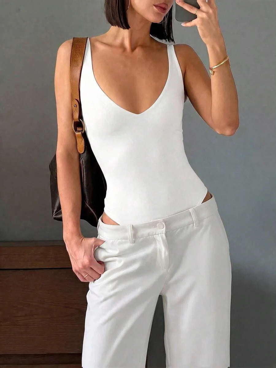 A woman wears a white deep V-neck bodysuit tucked into white trousers while holding a phone and carrying a brown leather bag.