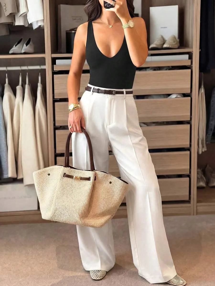 A woman in a closet takes a mirror selfie wearing a black V-neck bodysuit, white wide-leg trousers, and holding a woven straw tote bag.