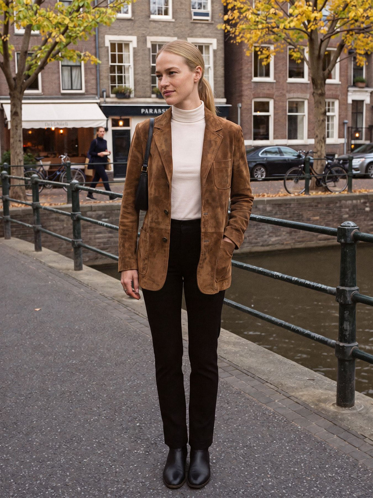Vrouw draagt de bruine jas in suèdelook open over een crème coltrui en zwarte pantalon aan een gracht in Amsterdam | Boutique Moda