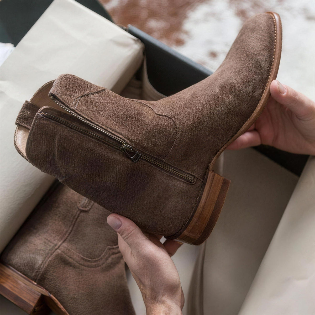 Bruine suède enkellaars met rits, vastgehouden boven schoenendoos | Boutique Moda