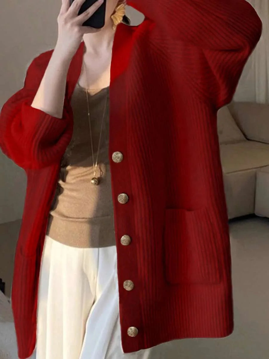 A woman wears an oversized red ribbed knit cardigan with gold buttons and front pockets, holding a phone up to her face.