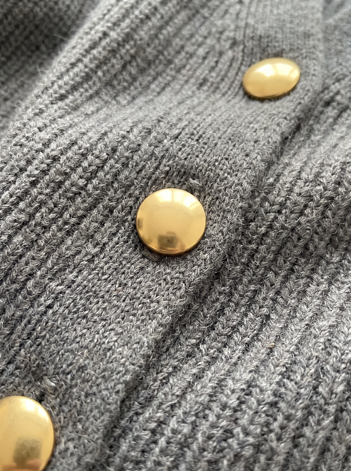 A woman wears an oversized grey knit cardigan with gold buttons and front pockets, holding a phone up to her face, close-up view