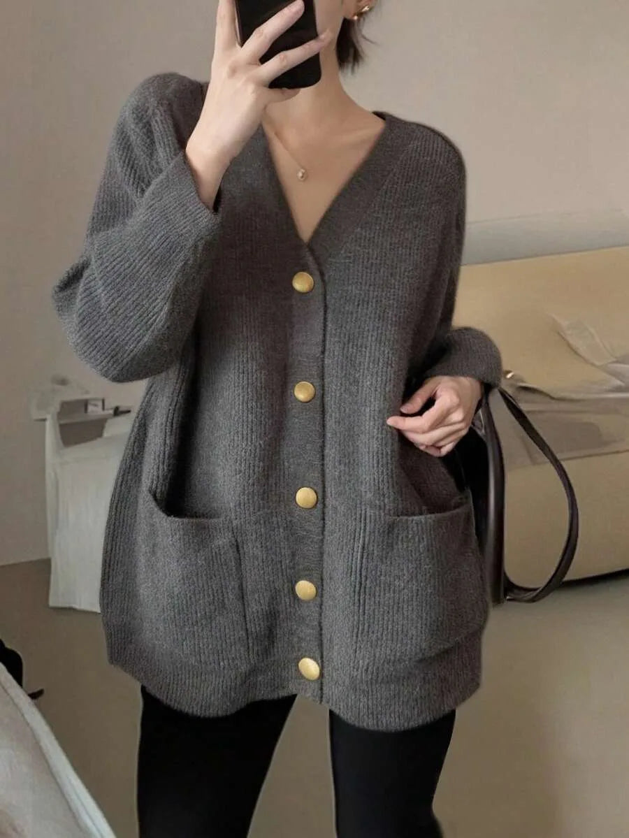 A woman wears a grey oversized ribbed cardigan with gold buttons and patch pockets, taking a mirror selfie with a phone covering her face.