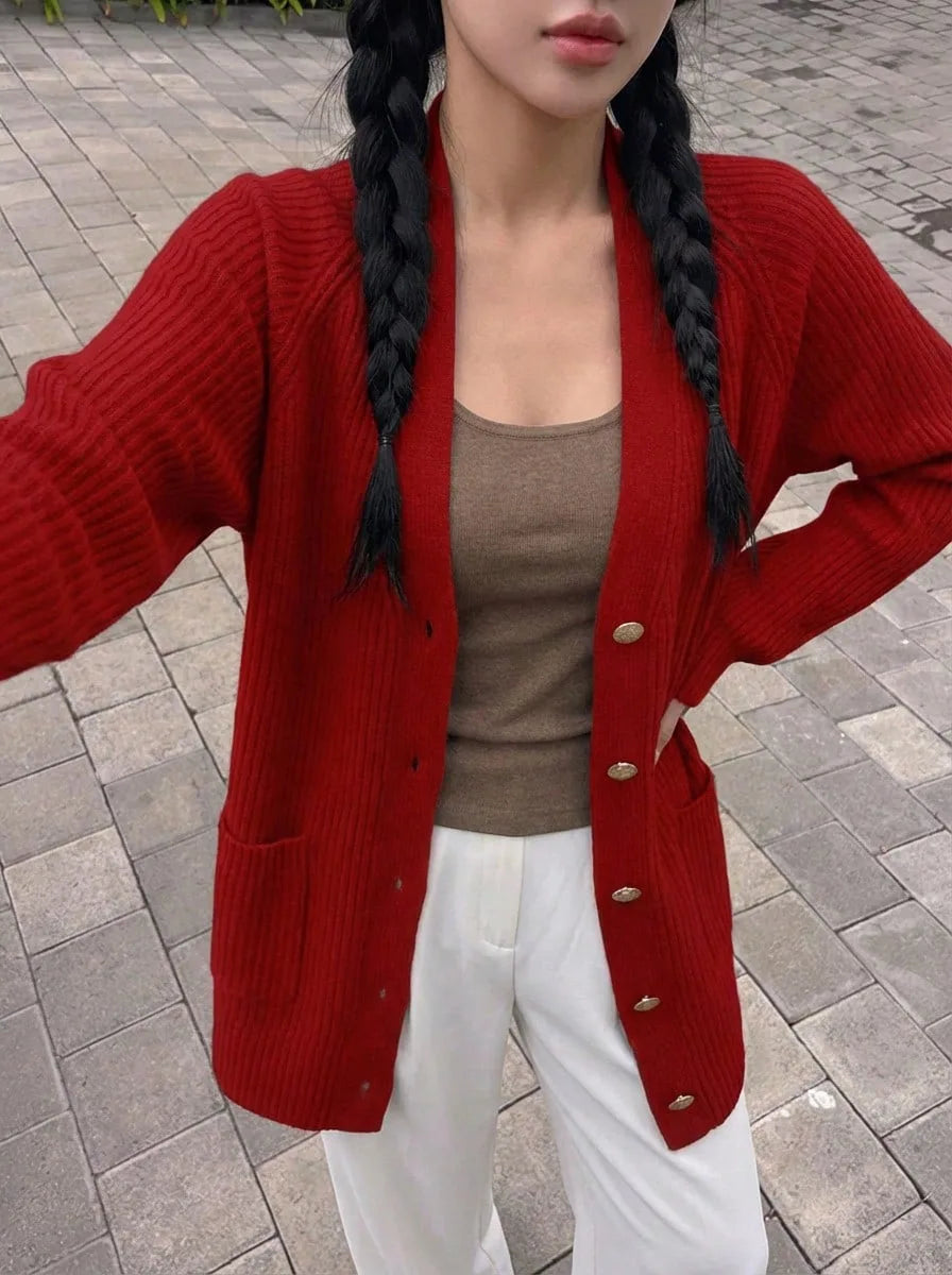 A woman wearing a red ribbed knit cardigan with gold buttons over a brown tank top, paired with white trousers, standing on a stone-paved street.