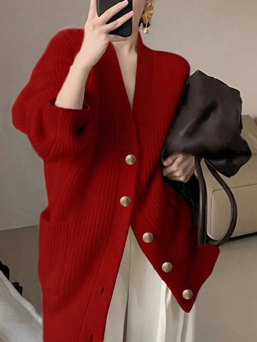 A person wears an oversized red ribbed knit cardigan with gold buttons while holding a phone and a black bag.