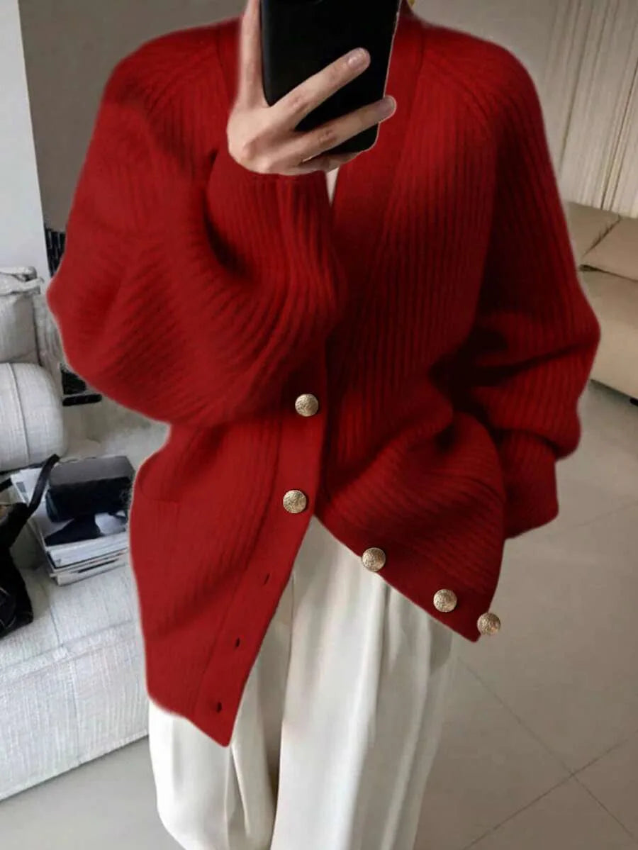 A person wears an oversized red ribbed knit cardigan with gold buttons, holding a phone to take a mirror selfie, paired with white wide leg trousers.
