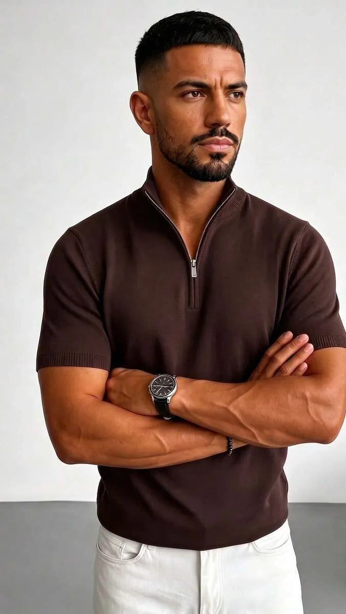 A man with arms crossed wears a brown short sleeve knit polo shirt with a half zip collar, paired with white pants.