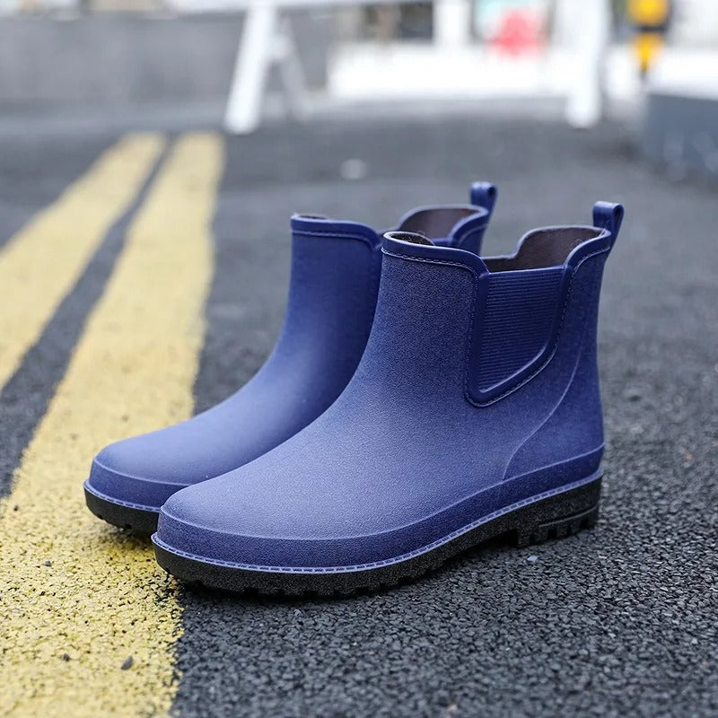 A pair of navy blue Chelsea style rubber rain boots with elastic side panels and black soles, standing on an asphalt road with yellow lane markings.