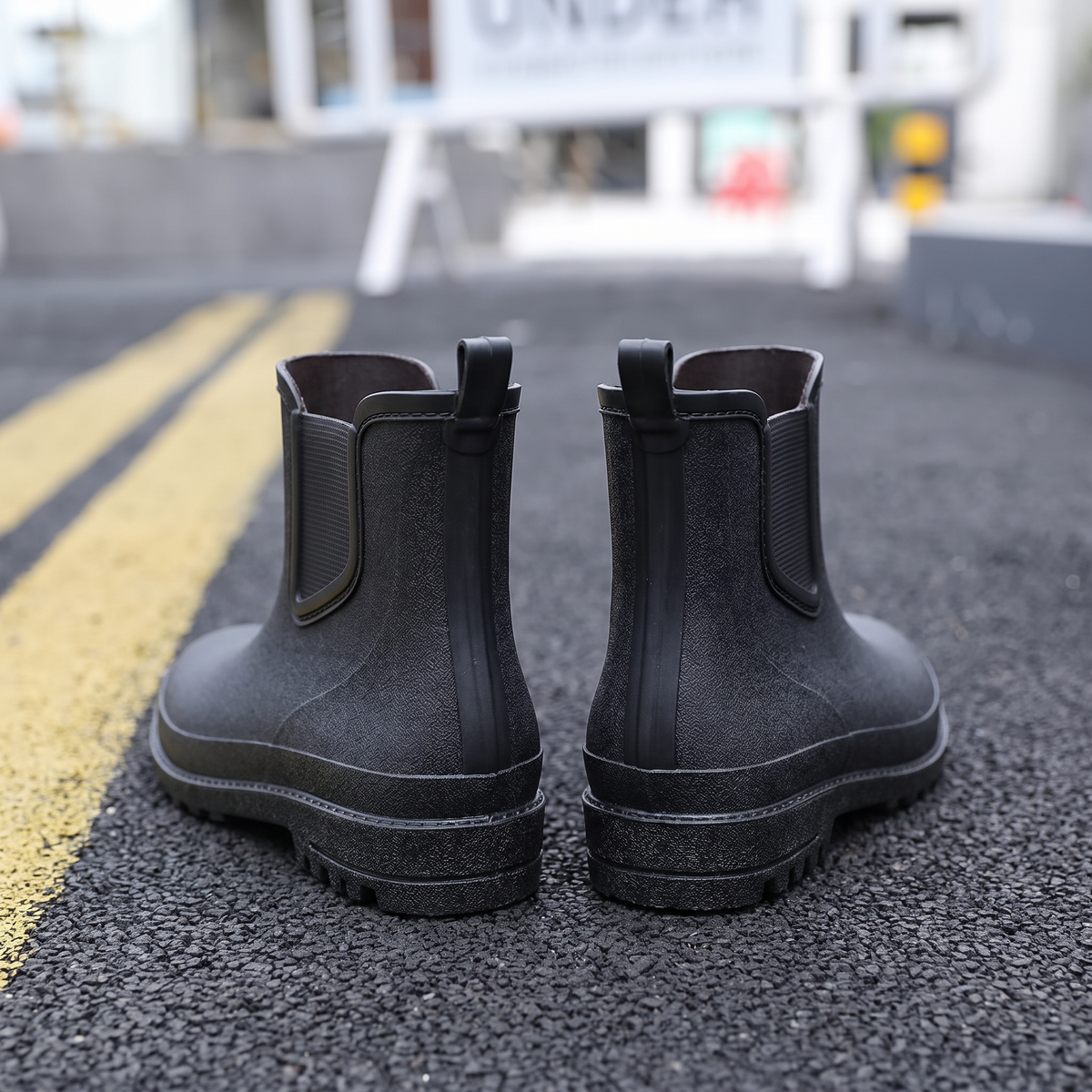 A pair of black ankle-height Chelsea style rubber rain boots with elastic side panels and pull tabs, standing on asphalt next to yellow road lines with a blurred street scene behind, back view