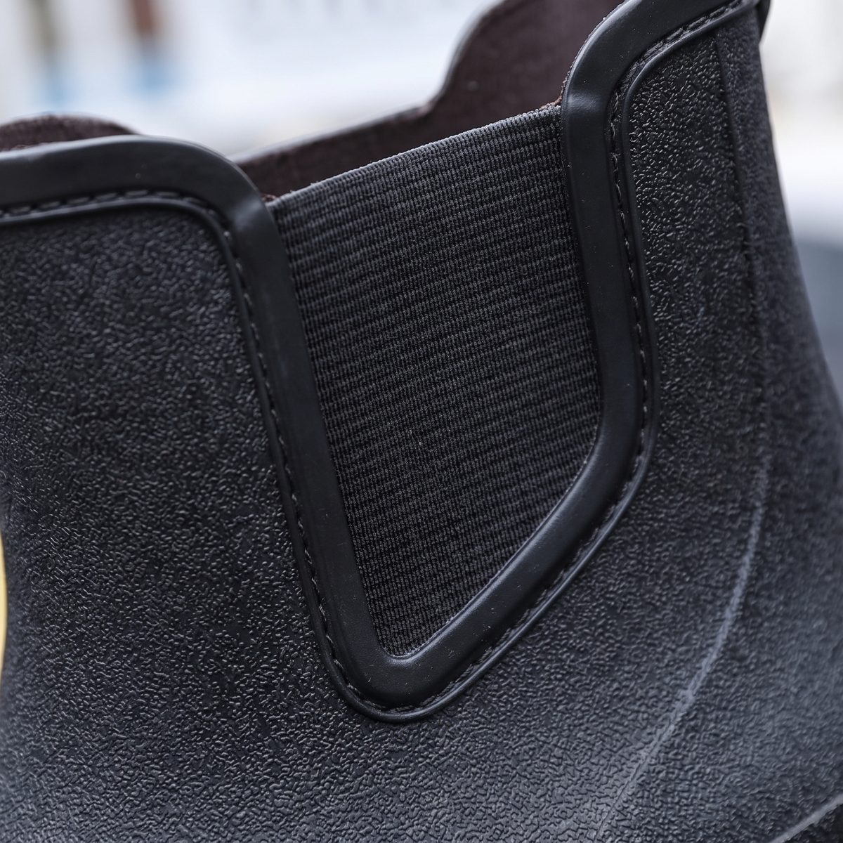A pair of black ankle-height Chelsea style rubber rain boots with elastic side panels and pull tabs, standing on asphalt next to yellow road lines with a blurred street scene behind, close-up view