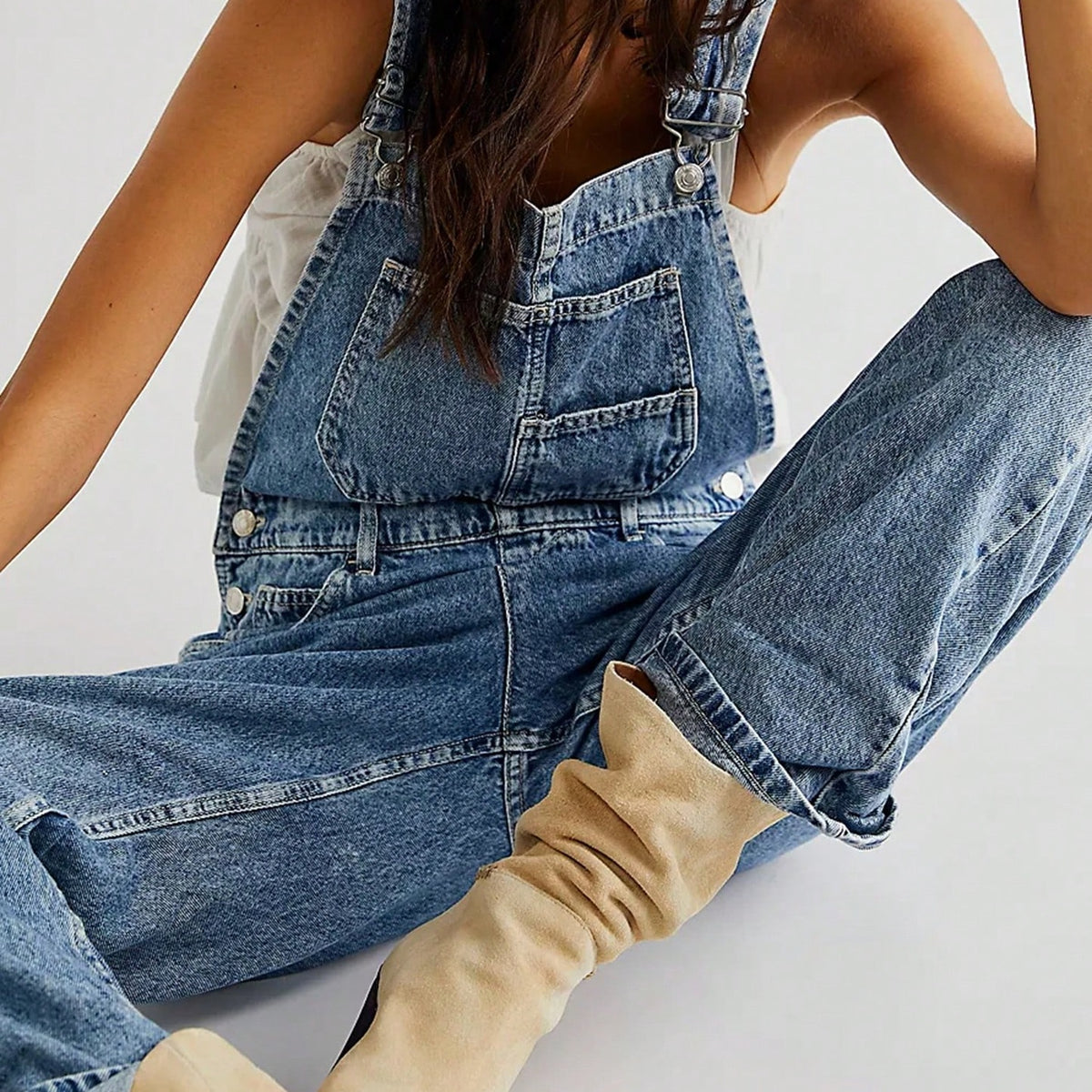 Blauwe denim overall los model, gedragen door zittende vrouw met beige laarzen | Boutique Moda