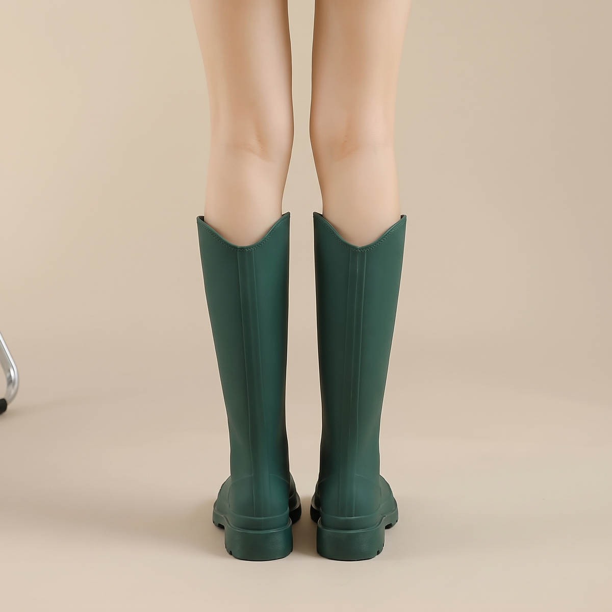 A woman wearing dark green tall rain boots with a chunky platform sole, shown from the knees down, back view