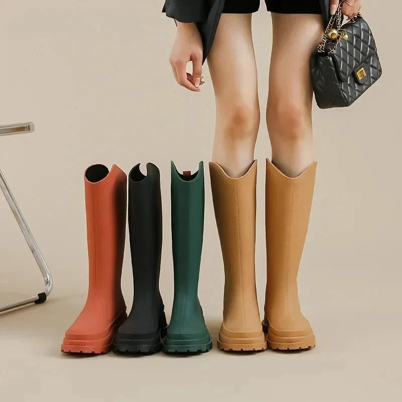 A woman's legs stand beside four pairs of tall knee high rain boots in orange, black, green and tan, with her carrying a quilted black handbag.