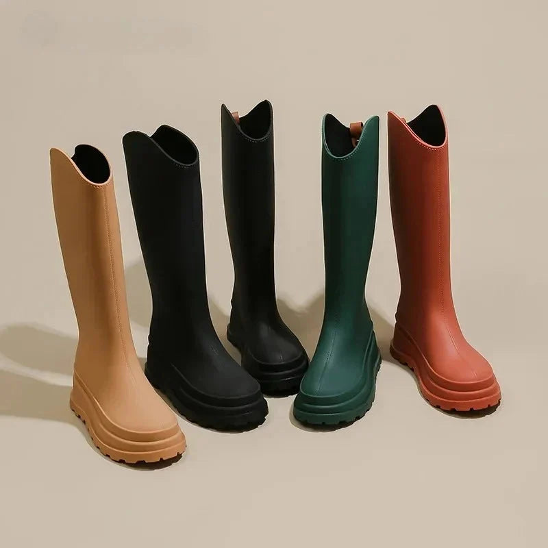 Five pairs of tall platform rain boots in camel, black, black, green and rust displayed standing on a plain beige background.