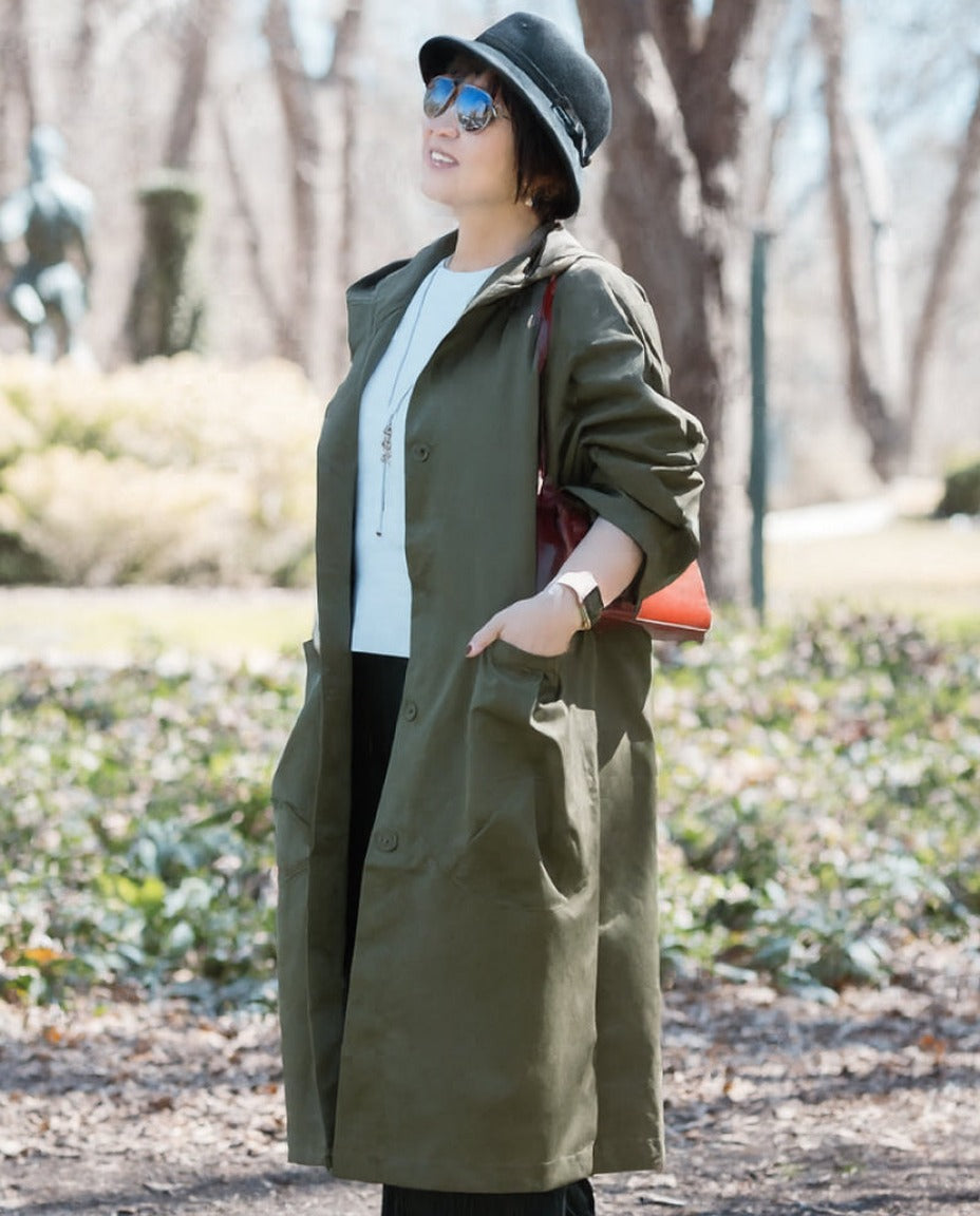 Vrouw draagt lange olijfgroene regenjas met hoed en zonnebril buiten | Boutique Moda
