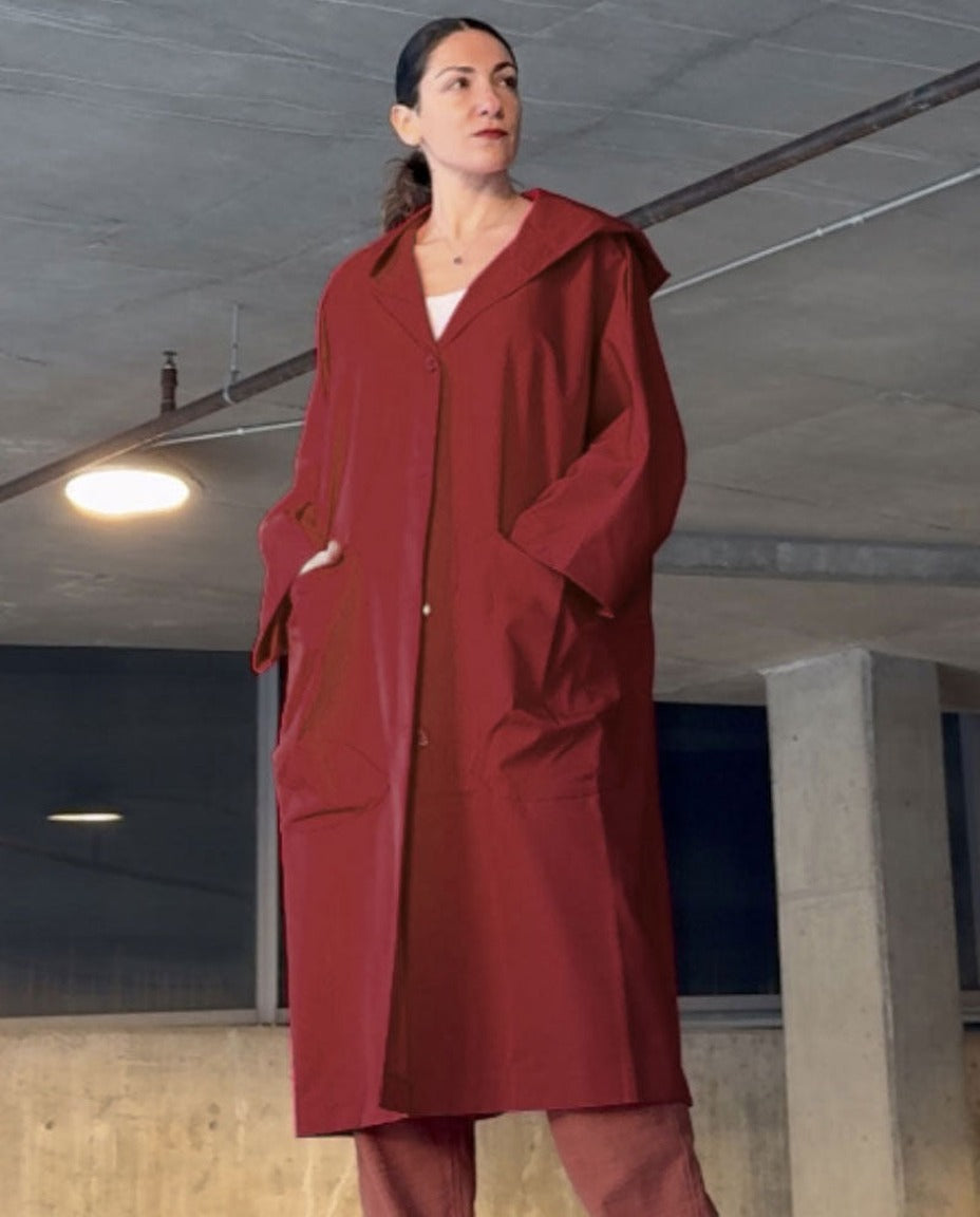 Vrouw draagt lange donkerrode oversized regenjas met capuchon, in parkeergarage | Boutique Moda