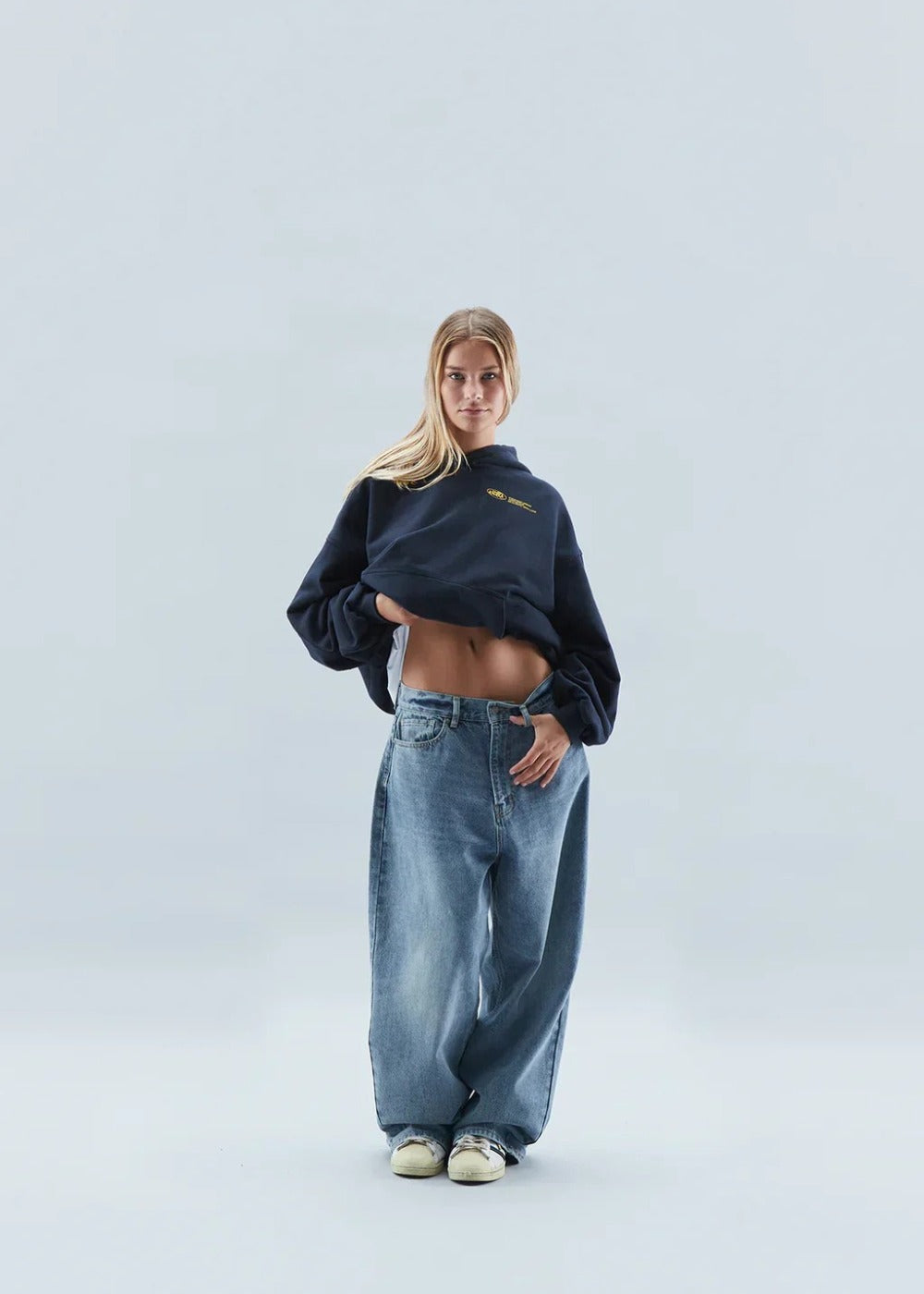Vrouw in een donkere oversized hoodie met een kleine gele print op de borst en een wijde blauwe spijkerbroek | Boutique Moda
