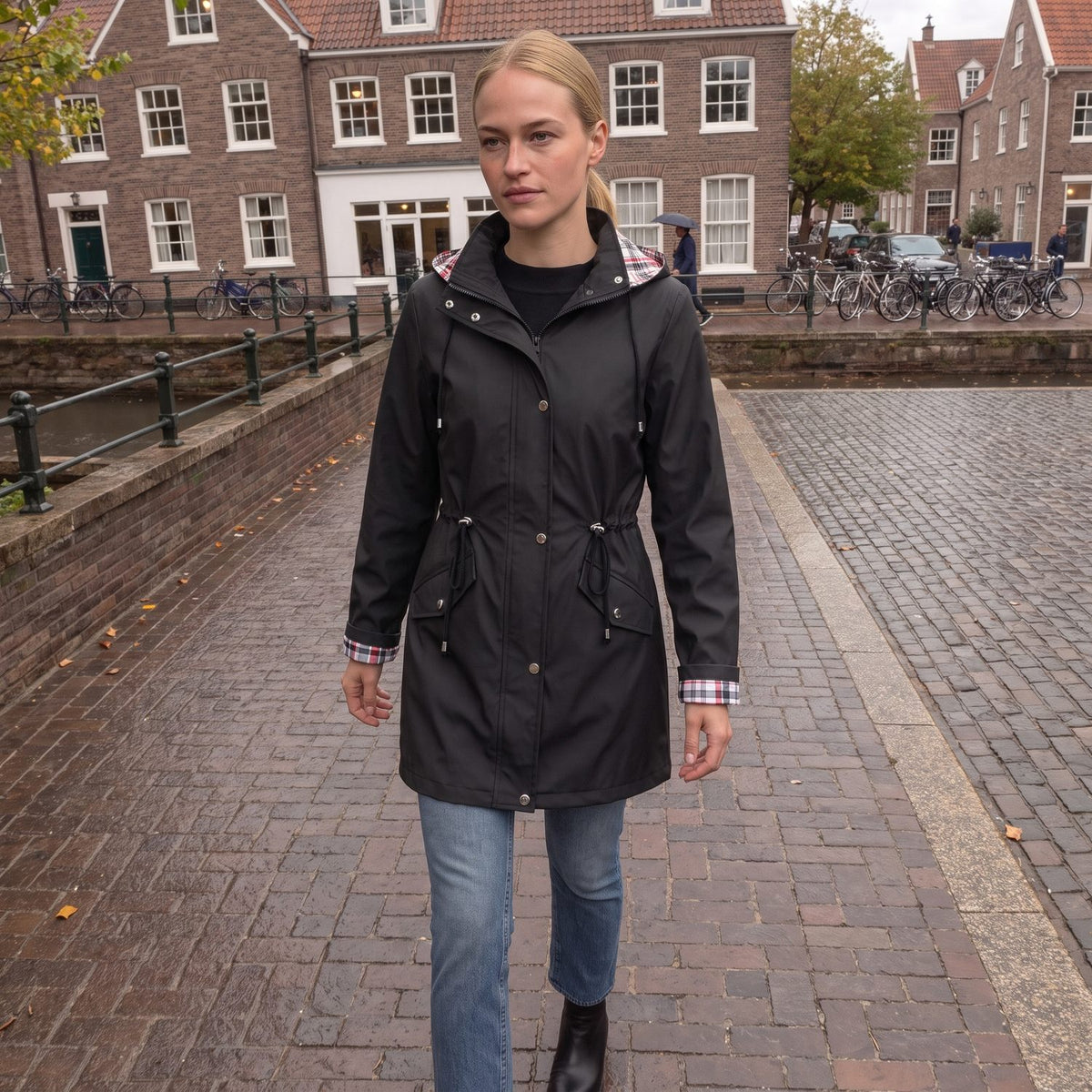 Vrouw in de zwarte regenjas met geruite capuchon aan een Nederlandse gracht