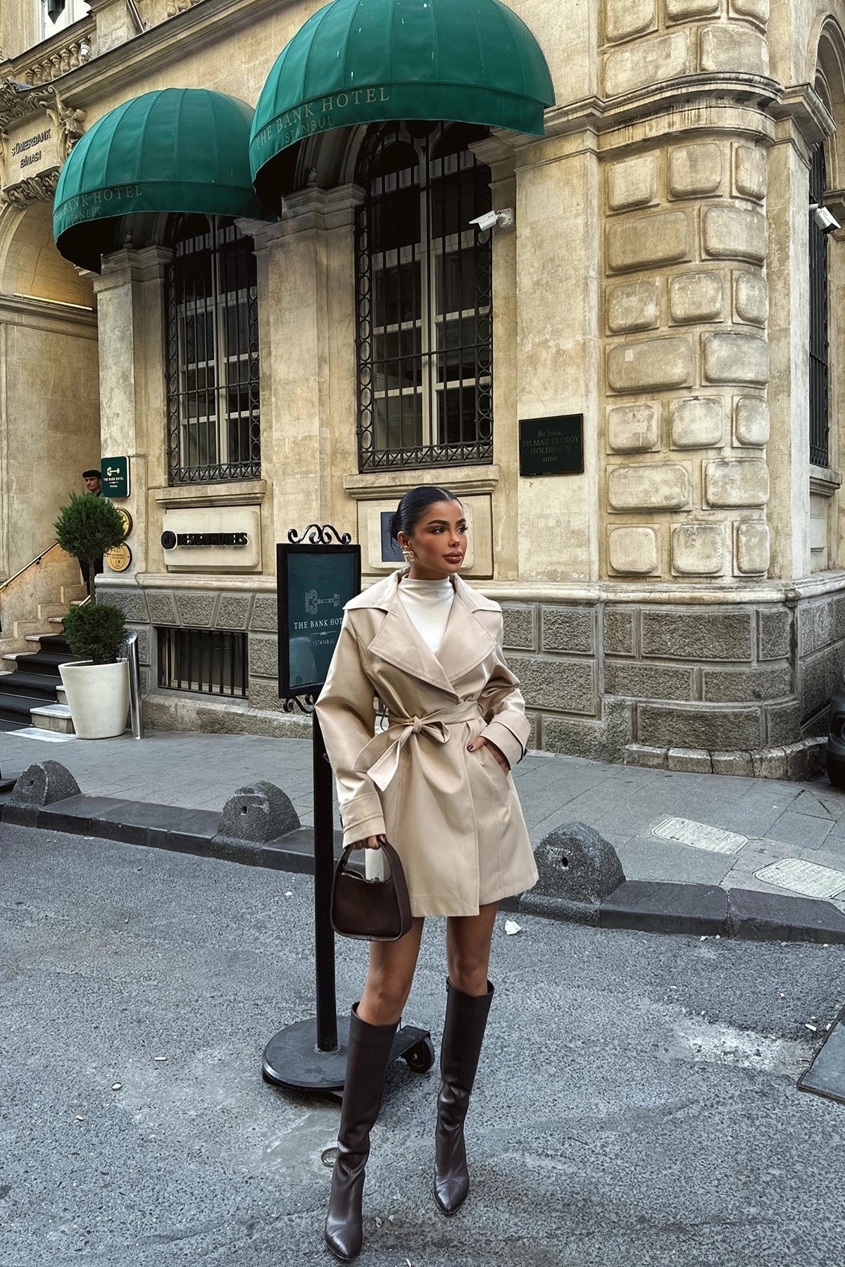 Vrouw in een korte beige trenchcoat met strikceintuur, gedragen met kniehoge laarzen op straat | Boutique Moda