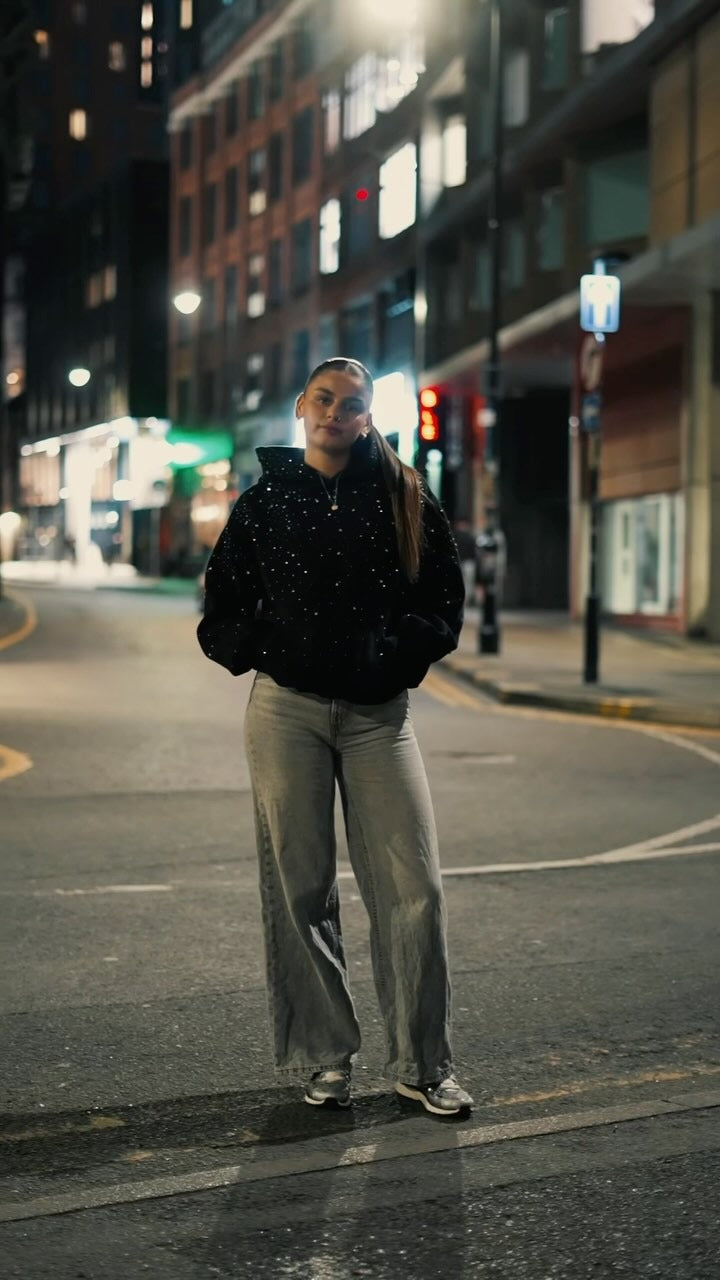 Vrouw in de zwarte unisex hoodie met strass en een lichte jeans, 's avonds op straat