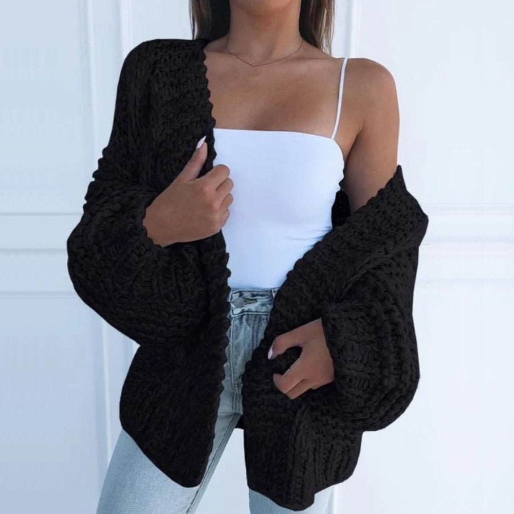Grofgebreid oversized damesvest in zwart, open gedragen over een wit topje, voor een witte muur met lijstwerk