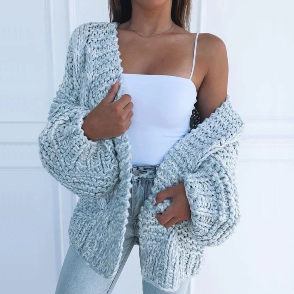 Grofgebreid oversized damesvest in lichtblauw, open gedragen over een wit topje, voor een witte muur met lijstwerk